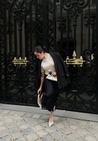 Cappotto nero su un maglione beige, gonna nera con spacchi e tacchi nudi. Tiene una pochette bianca, in piedi accanto a un cancello nero ornato.