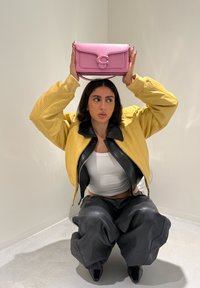 Femme accroupie contre un mur beige, portant une veste jaune et un pantalon noir, tenant un sac à main rose au-dessus de sa tête avec les deux mains.