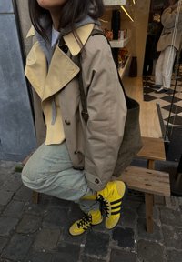 Veste beige et jaune avec un large col, sweat à capuche gris en dessous, pantalon bleu clair, et chaussures Adidas jaune vif à rayures noires.