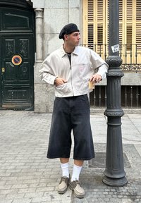 Gaišpelēka jaku ar rāvējslēdzēju un priekšējo kabatu, melnas džinsus līdz ceļgaliem, baltas zeķes un bēšas kurpes, stāv uz ķieģeļu ielas blakus laternai.