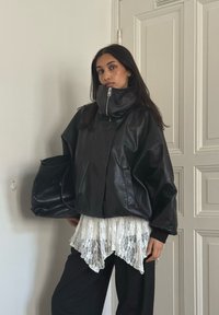 Svart skinnjakke i stor størrelse med høy krage, glidelås foran og senkede skuldre. Kombinert med en hvit blonde-topp og svarte bukser.