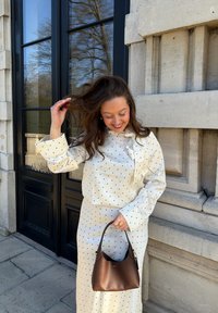 Femme souriante touchant ses cheveux, portant une robe blanche à pois, tenant un sac à main en cuir marron, debout près d'un bâtiment en pierre.