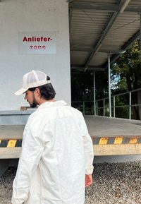 Bela majica s dolgimi rokavi, z vidnimi šivi, oblečena z svetlo barvno kapo, stoji ob območju za nalaganje z opozorilnim pasom na robu.