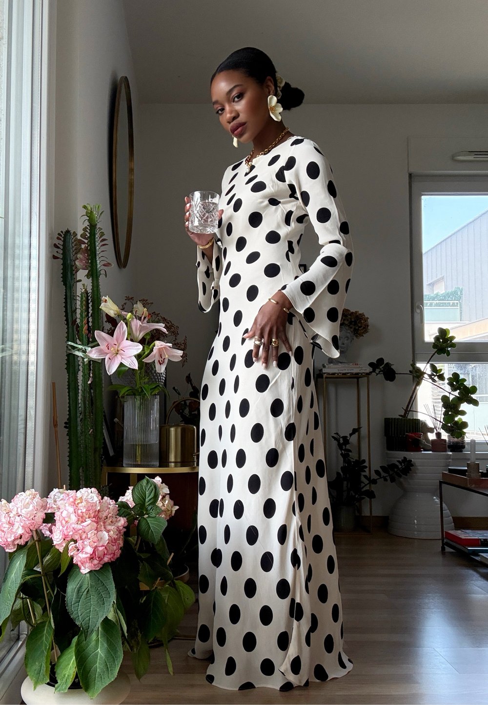 Lange, vloeiende jurk in roomkleur met grote zwarte stippen. Klokmouwen, soepele stof en gelaagde bloemaccenten op de voorgrond.