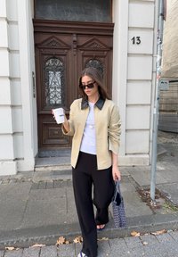 Jachetă bej cu fermoar și guler negru, tricou alb, pantaloni negri și papuci albastre. Ținând o cană de cafea și o geantă cu model.