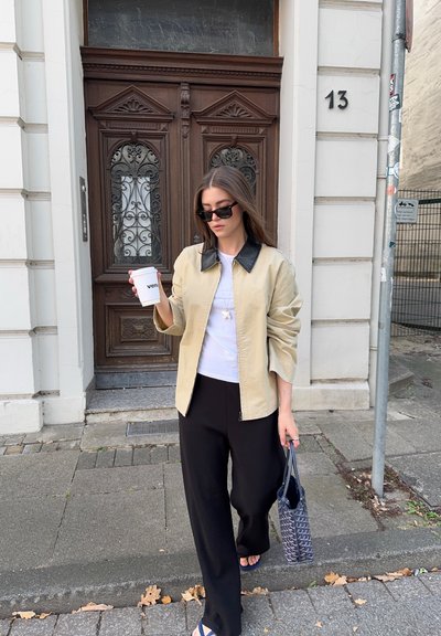 Chaqueta beige con cremallera y cuello negro, camiseta blanca, pantalones negros y chanclas azules. Sosteniendo una taza de café y un bolso de estampado.