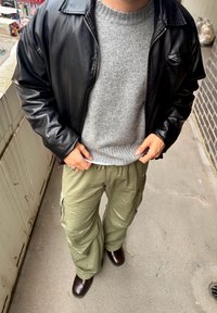 Fekete bőrdzseki, szürke kötött pulóver, oliva zöld katonai nadrág és barna csizmák. Részletek: bordázott mandzsetták és több zseb.