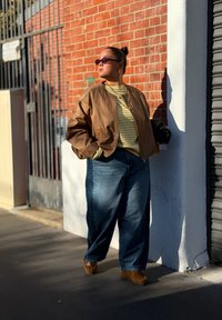 Brun bomberjakke, gulstripete skjorte, blå baggy jeans, brune mokkasinsko. Tilbehør: runde solbriller. Bakgrunn: murvegg.