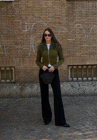 Cardigan verde măsliniu cu dungi, cu guler de blană, pantaloni negri cu picior larg și o geantă mică neagră, pe un fundal de zid de cărămidă texturată.