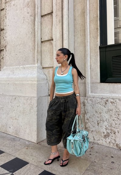 Joven mujer con coleta que lleva una camiseta sin mangas azul, pantalones anchos de camuflaje, tacones negros, joyas plateadas, sosteniendo un gran bolso azul claro junto a un muro de piedra.