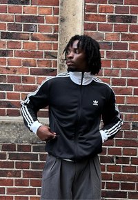 Černá bunda na zip s bílými pruhy podél rukávů, spárovaná se šedými, plisovanými kalhotami. V pozadí texturovaná cihlová zeď.