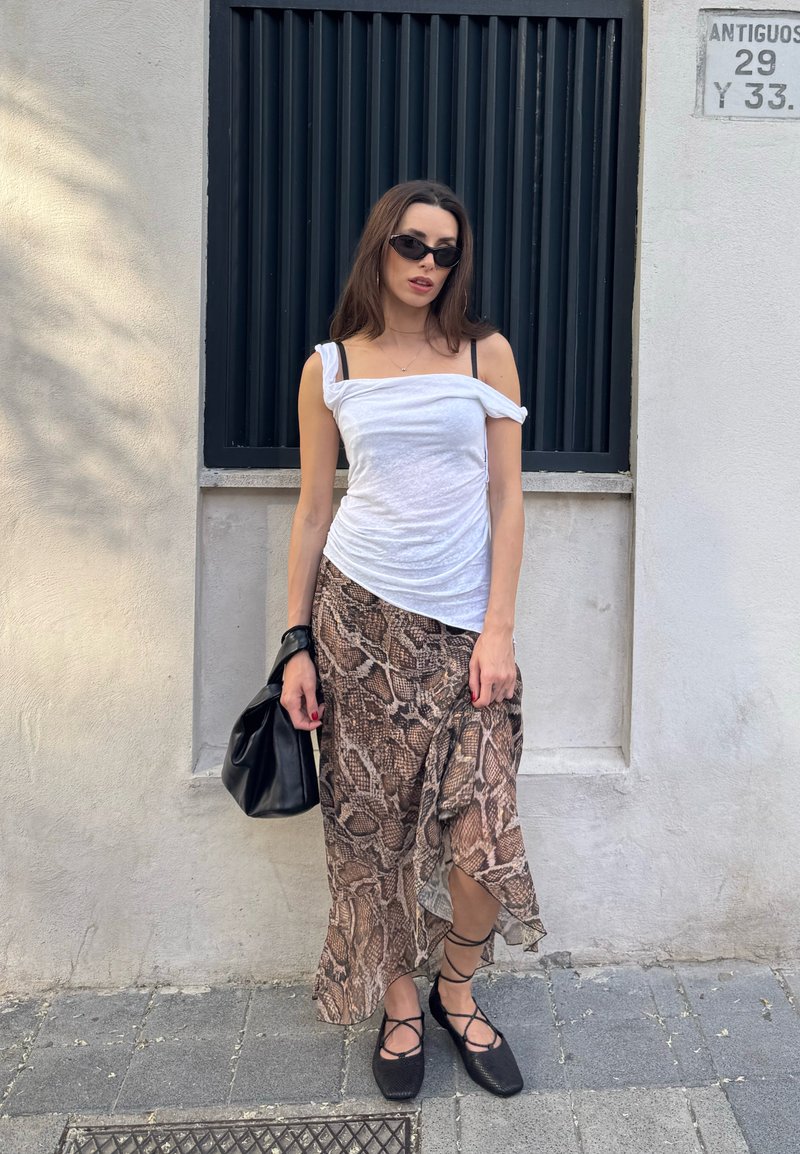 Femme portant un top blanc à épaules dénudées, une jupe imprimée serpent, des ballerines noires à lacets, des lunettes de soleil noires, tenant un sac à main noir, debout près d'un mur avec des barreaux aux fenêtres.