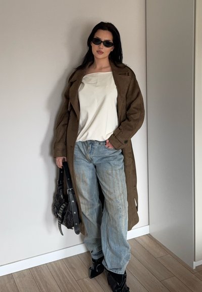 Mujer con gafas de sol oscuras, abrigo largo marrón, top blanco, vaqueros azules holgados, sosteniendo un bolso negro, de pie contra una pared clara y lisa.