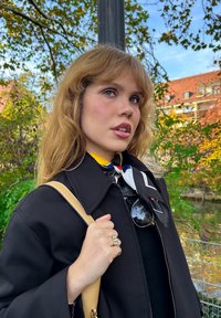 Veste noire avec un col, écharpe jaune à rayures et sac à épaule beige. Cheveux longs et ondulés avec une texture naturelle. Des lunettes de soleil complètent le look.