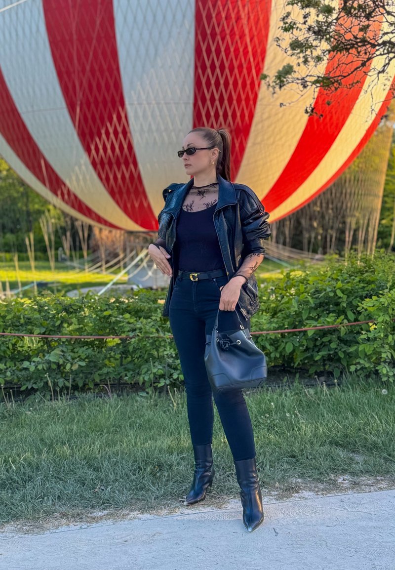 Kvinde iført sort læderjakke, sort top, bukser, støvler og solbriller står og holder en sort håndtaske foran en stor rød- og hvidstribet ballon.