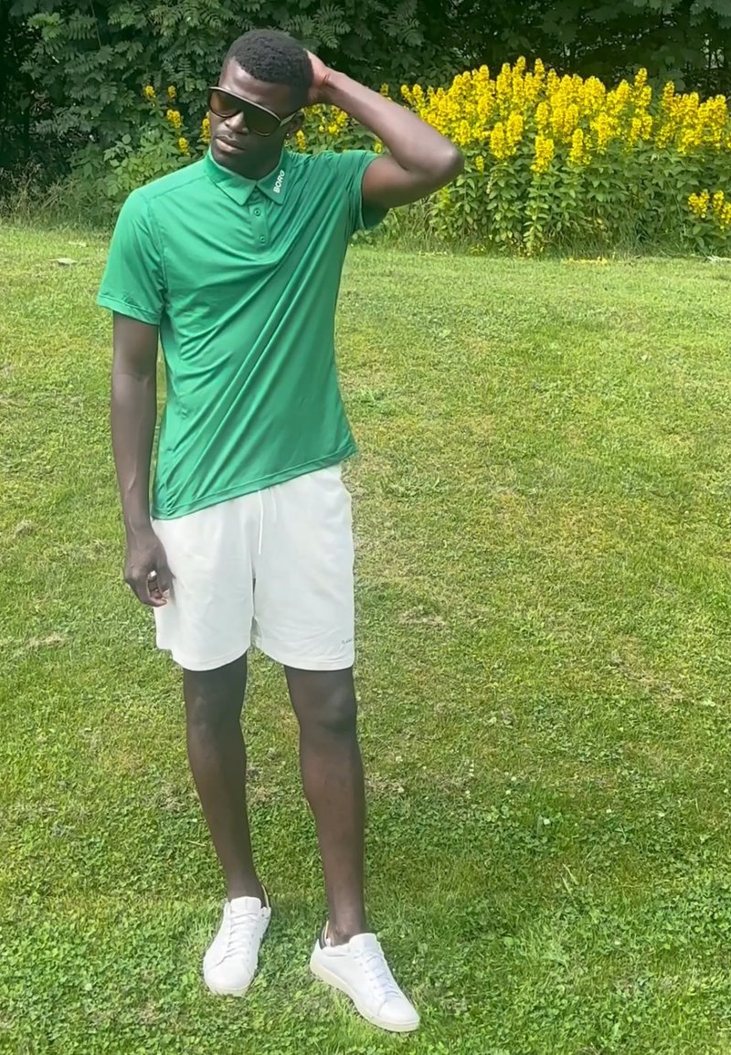 Homme en polo vert, short blanc et lunettes de soleil, debout sur l'herbe avec une main sur la tête près de plantes jaunes en fleurs.