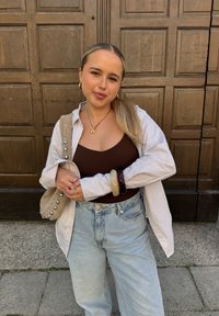 Weißes und gestreiftes Oversized-Hemd, braunes enges Top, hochgeschnittene hellblaue Jeans, beiger Bag mit silbernen Nieten, geschichtete Armbänder, Holz-Hintergrund.