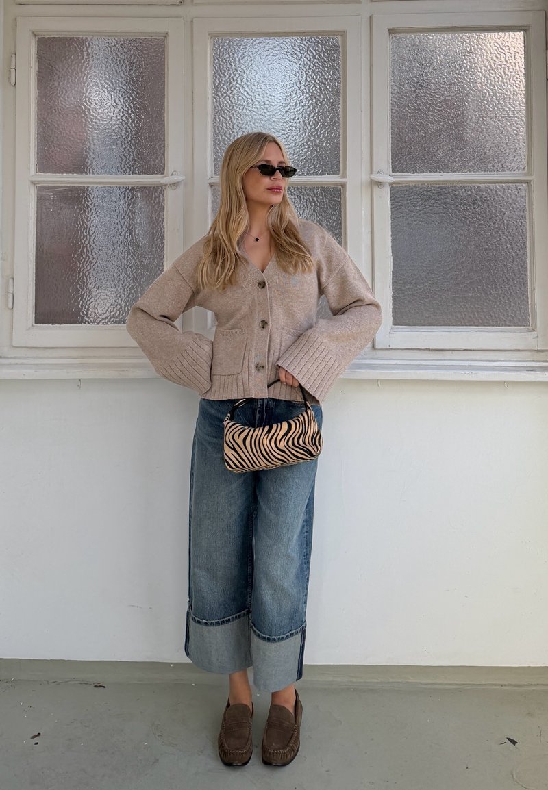 Γυναίκα με μακριά ξανθά μαλλιά που φοράει γυαλιά ηλίου, μπεζ ζακέτα, φαρδιά τζιν με μανσέτες, καφέ μοκασίνια και κρατά τσάντα με ρίγες ζέβρας σε εσωτερικό χώρο.