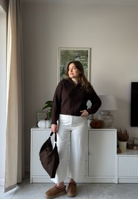 Brązowy prążkowany sweter na zamek, białe szerokie spodnie, brązowa teksturowana torebka oraz brązowe kapcie z jasną podeszwą, stojące obok szafki.