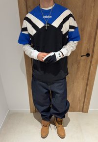 Personne portant un t-shirt Adidas bleu, blanc et noir, des gants blancs et noirs, un jean large, des bottes brunes et un collier avec une croix, debout à l'intérieur.