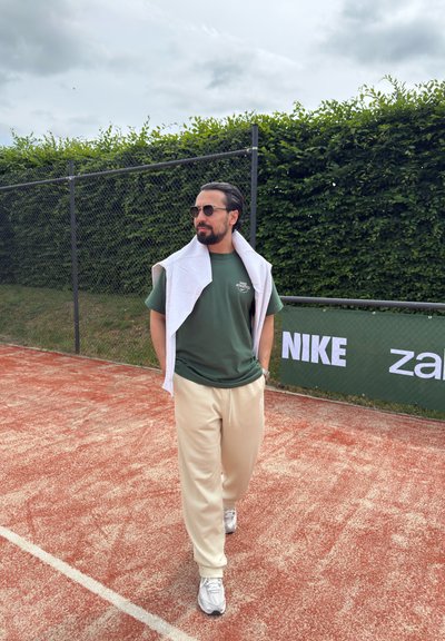 Hombre con gafas de sol, camiseta verde de Nike, sudadera blanca sobre los hombros y pantalones beige camina por una pista de tenis de tierra batida cerca de una valla con un cartel de Nike.