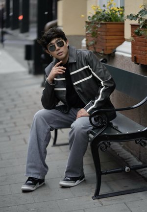 Chaqueta de cuero negra con rayas blancas, camiseta negra, pantalones grises y zapatillas negras. Sentado en un banco con flores en macetas de fondo.