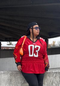 Červený dres s čiernymi a oranžovými akcentmi, s potlačou "03", doplnený čiernymi nohavicami a malou oranžovou taškou na rameno. Zložité zapletené účes a slnečné okuliare dotvárajú vzhľad.