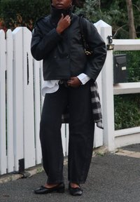 Svart skinnjakke med dobbelspent knapping, hvit skjorte, rutet flanell, grå vidbente jeans og svarte flate sko. Står mot et hvitt gjerde.