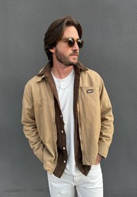 Brązowa kurtka z sztruksu z zewnętrzną warstwą w kolorze khaki, pod spodem biała koszulka, okulary przeciwsłoneczne; gładka faktura i luźny krój.