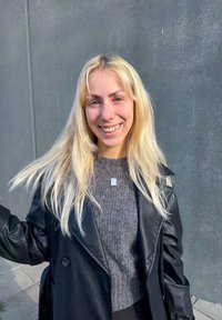 Sort læder dobbeltknappet frakke over en grå struktureret sweater. Personen har langt blondt hår og bærer en sølv firkantet vedhængs halskæde.