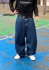 Weite dunkle Denim-Hose mit einem fünf-Knopf-Verschluss, großen Taschen und weißen Nähten. Kombiniert mit einem schwarzen Langarmshirt und weißen Sneakers.