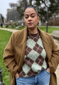 Manteau en laine marron porté sur un pull à motif losanges verts et marron ; boucles d'oreilles créoles dorées et chaîne en collier ; jean en denim bleu. Cadre extérieur.