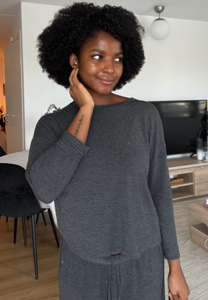 Joven con peinado afro, vestida con top y pantalones grises de manga larga, se toca la cara mientras está de pie en una sala de estar moderna con televisión y muebles de madera.
