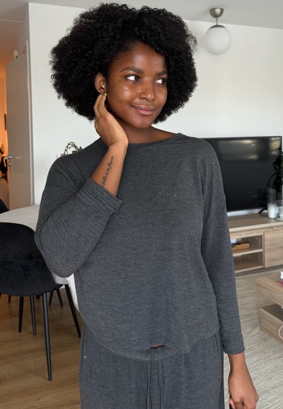 Joven con peinado afro, vestida con top y pantalones grises de manga larga, se toca la cara mientras está de pie en una sala de estar moderna con televisión y muebles de madera.