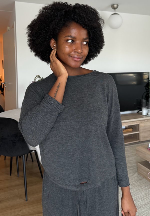 Jeune femme aux cheveux afro portant un haut à manches longues gris et un pantalon, touchant son visage, debout dans un salon moderne avec une télévision et des meubles en bois.
