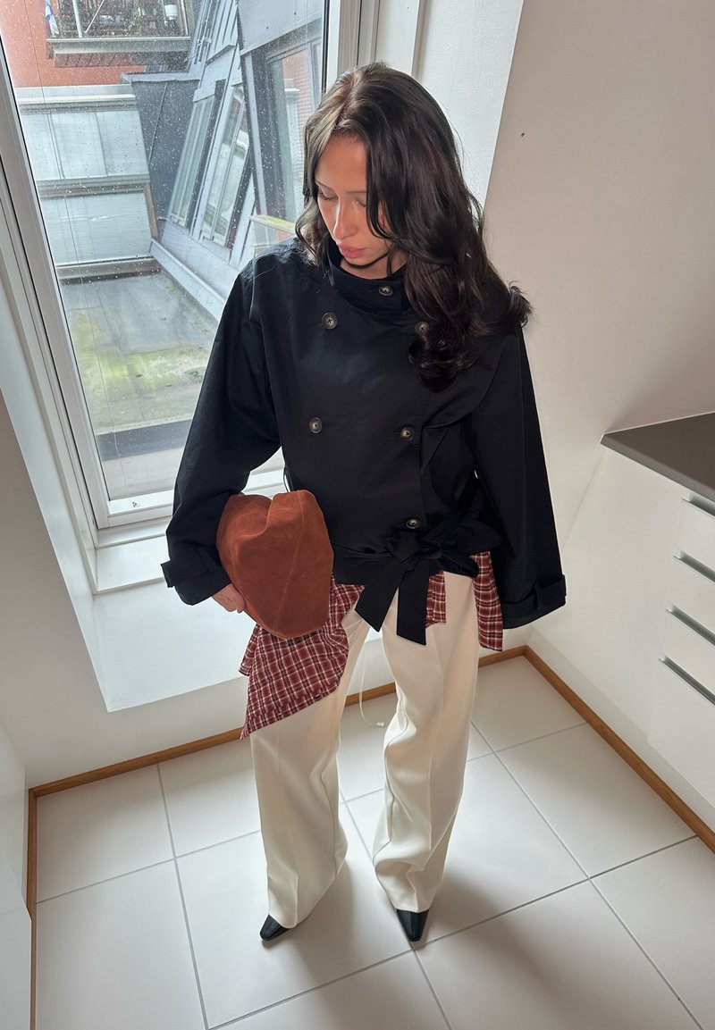 Black double-breasted jacket with wide sleeves, red plaid accent underneath, white wide-leg trousers, and a brown fabric hat.