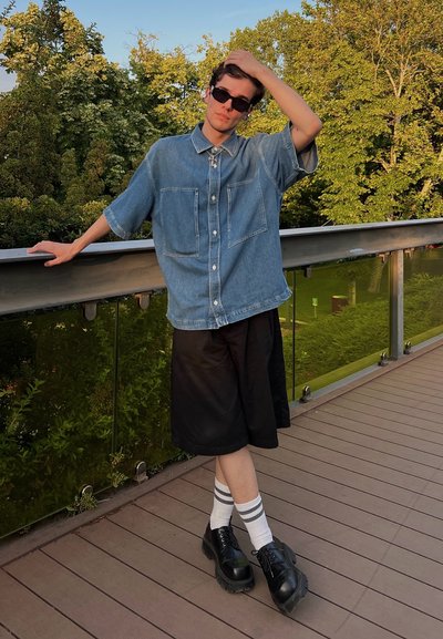 Camisa de mezclilla con botones y dos bolsillos frontales, pantalones cortos negros hasta la rodilla y zapatos de plataforma negros. Medias blancas con rayas grises visibles.