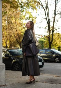 Długi ciemno-brązowy skórzany płaszcz z frędzlami na ramionach, w zestawieniu z ciemną torebką i brązowymi szpilkami, na tle miejskiego jesiennego krajobrazu.