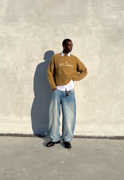 Suéter canela de punto con el texto "Cena Tarde", llevado sobre una camisa blanca. Jeans oversized azul claro y zapatos negros. Fondo de concreto.