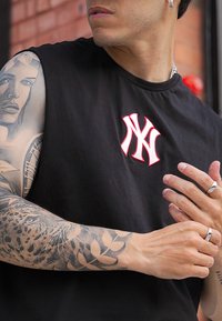 Črna majica brez rokavov z belim NY logotipom obrobljenim v rdeči barvi. Oseba nosi podrobne tetovaže na mišičnih rokah. Na prstih nosi srebrne prstane.