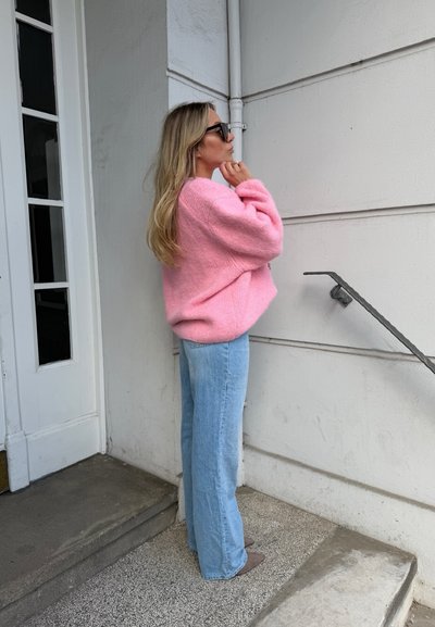 Mujer con suéter rosa y vaqueros azules está junto a una pared blanca cerca de unas escaleras, ajustándose las gafas de sol con la mano derecha.