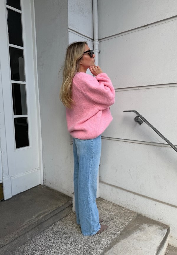 Femeie cu pulover roz și blugi albaștri stă lângă un perete alb aproape de scări, ajustând ochelarii de soare cu mâna dreaptă.