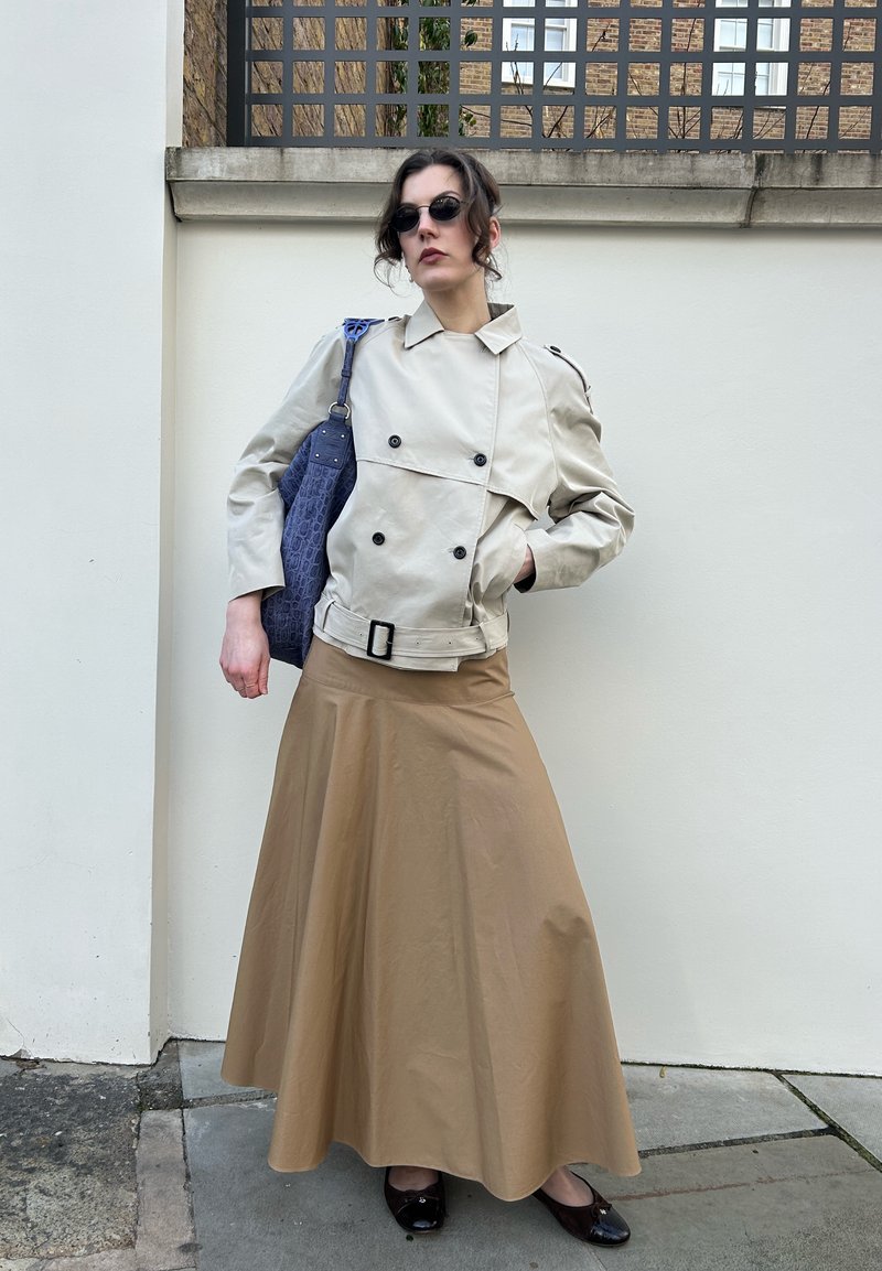 Femme portant une veste beige à double boutonnage et une longue jupe camel, portant un sac à bandoulière bleu texturé, debout contre un mur blanc avec une rampe en métal.