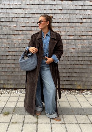 Abrigo de ante marrón sobre una camisa de mezclilla azul, jeans anchos azul y bolso tejido gris. Unas gafas de sol oscuras completan el look.