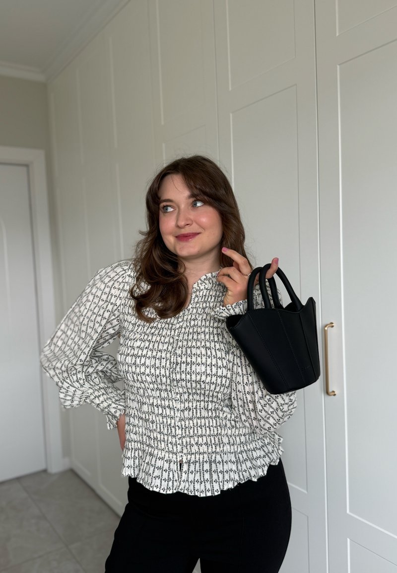 Femme aux cheveux bruns portant un chemisier à motifs et un pantalon noir tient un petit sac à main noir sur son épaule dans une pièce avec des armoires blanches.