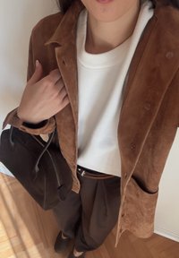 Brūns zamšādas žakete pār baltu džemperi, brūnas bikses, rokās tumša ādas soma. Raksturīgas mīkstas struktūras un kontrastējošas krāsas.