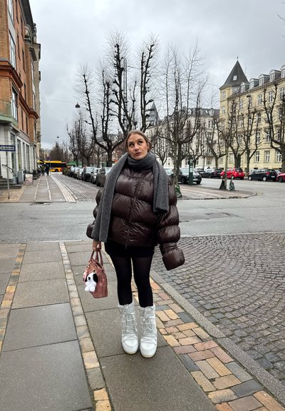 Chaqueta de plumas marrón, bufanda gris, mallas negras, botas de nieve blancas. Bolso rosa con un accesorio de peluche. Acera adoquinada y calle adornada de árboles.