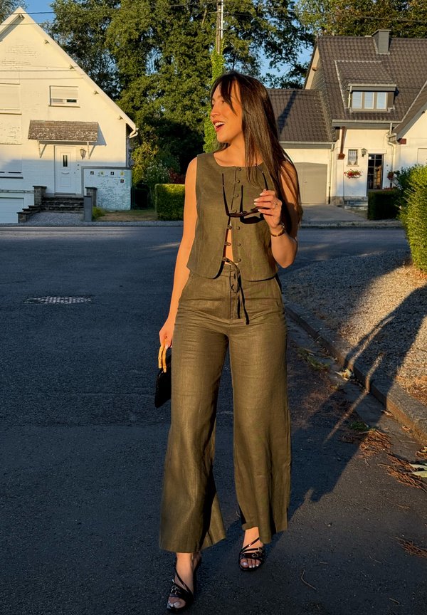 Donna sorridente che cammina per strada con top verde oliva senza maniche e pantaloni, tenendo occhiali da sole e una piccola borsa, vicino alle case sotto al sole.