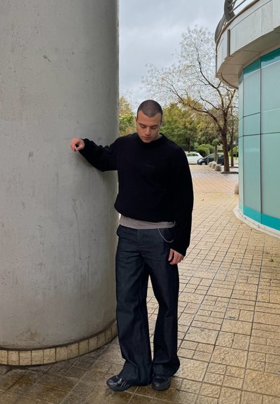 Suéter de punto negro, camiseta gris debajo, pantalones de mezclilla oscuros y zapatos negros. La persona se apoya contra un pilar de concreto en un entorno al aire libre.