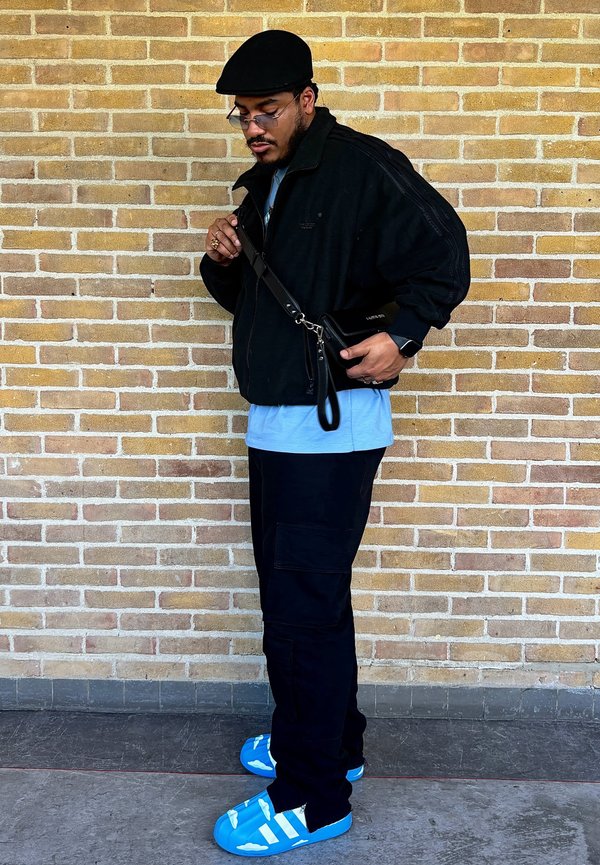 Casaco preto com fecho, t-shirt azul clara, calças cargo pretas, sapatos azuis com design de nuvens brancas. A segurar uma mala preta à tiracolo.